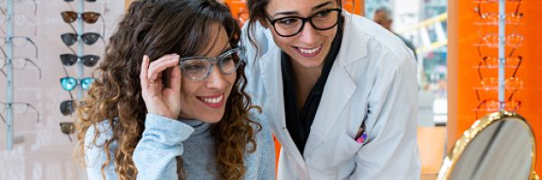 Happy female optician and client wearing glasses and looking at the mirror. Stand of glasses in the background